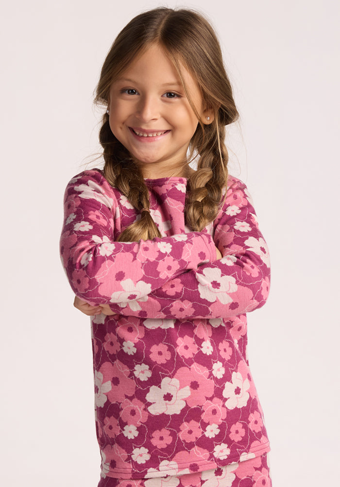 A young girl with long brown hair in braids smiles with arms crossed, wearing the Woolx Kids Explorer Base Layer Top - Rosie Posie, inspired by Merino wool base layers, against a plain light background. 