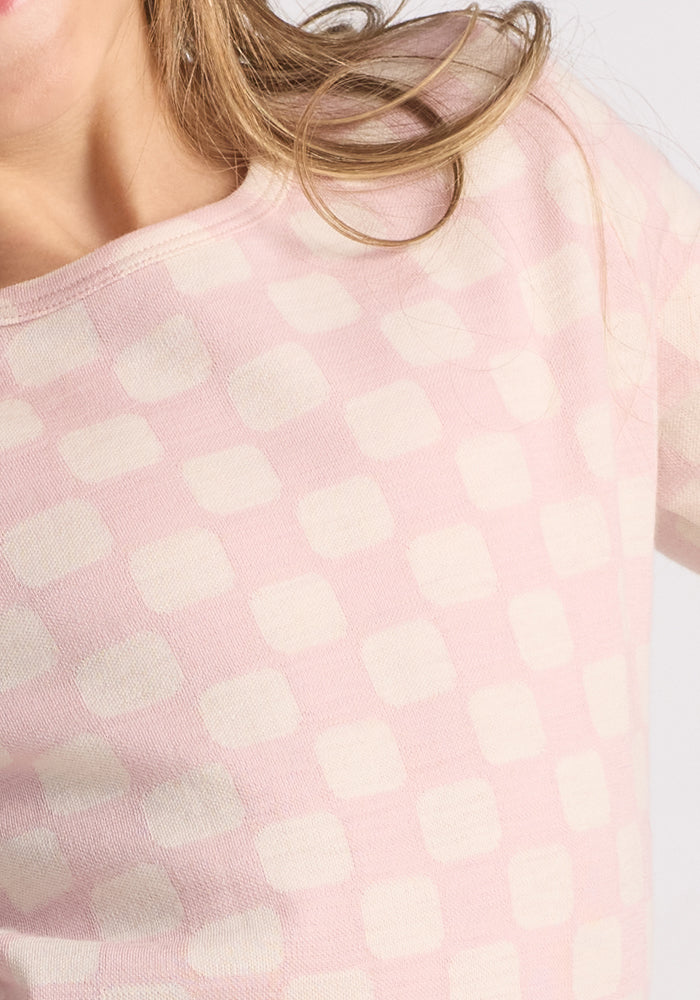 Close-up of a child wearing the Woolx Kids Explorer Base Layer Top in Hopscotch Pink, featuring off-white squares and made from soft Australian Merino wool. Part of their face, neck, and long light brown hair are visible against a plain background.
