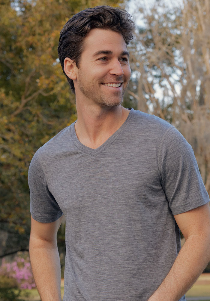 A man with dark hair and a short beard smiles outdoors, wearing the Woolx Ashton V Neck in Graphite Heather. Behind him, trees with green and bare branches are visible.