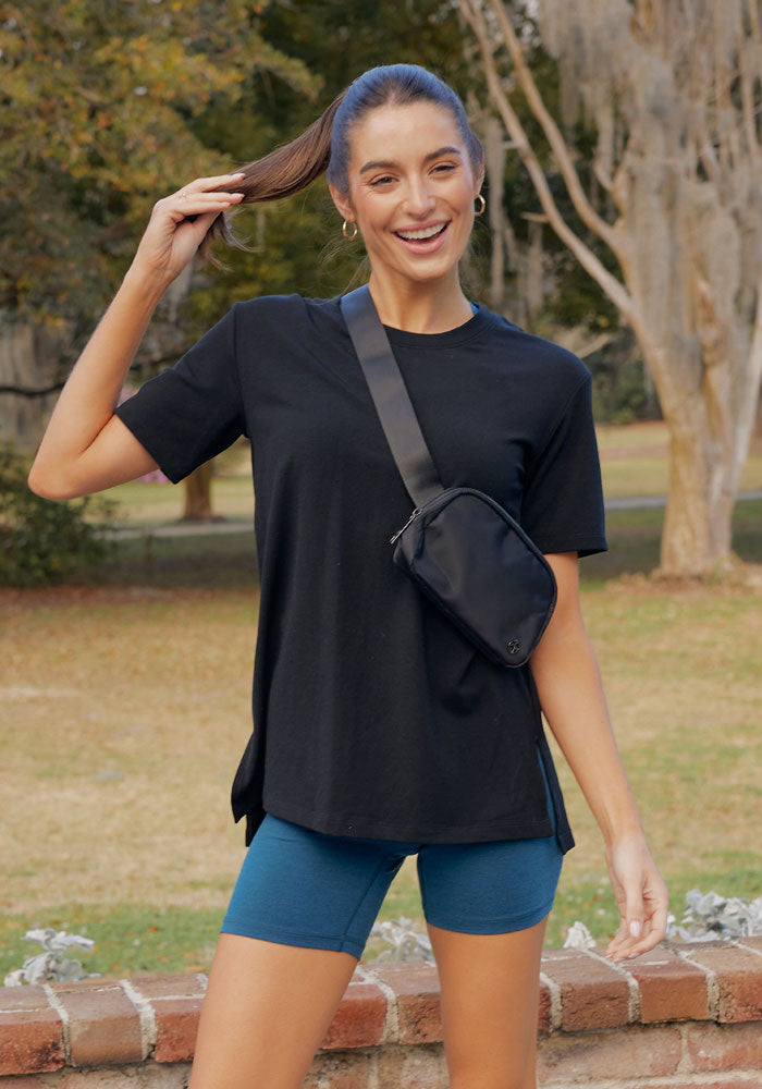 A woman outdoors smiles while holding her ponytail. She wears the Woolx Audrey Tee - Black, blue bike shorts, and a black crossbody bag. Trees and greenery are in the background.