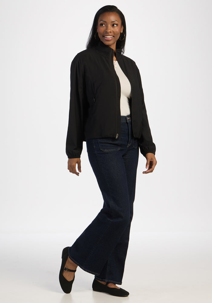Woman in Woolx Cooper Windbreaker - Black, white top, dark jeans, black flats, smiling against a white background.