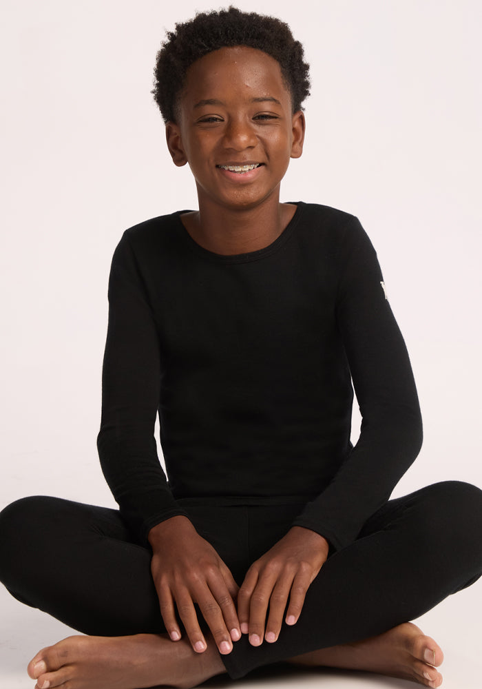 A smiling child with short curly hair sits cross-legged on the floor, wearing the Woolx Kids Explorer Base Layer Top in black, against a plain light background.