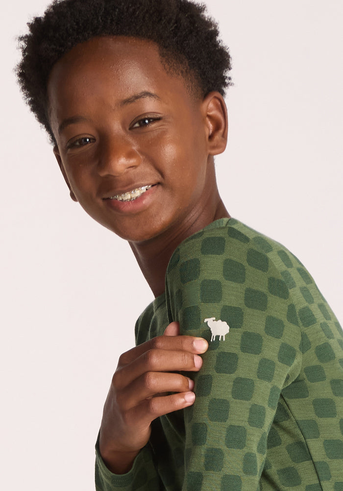 A smiling young boy with short curly hair models the Woolx Kids Explorer Base Layer Top - Frog Hop, touching the small white sheep logo on the sleeve to highlight its soft, breathable Merino wool comfort.