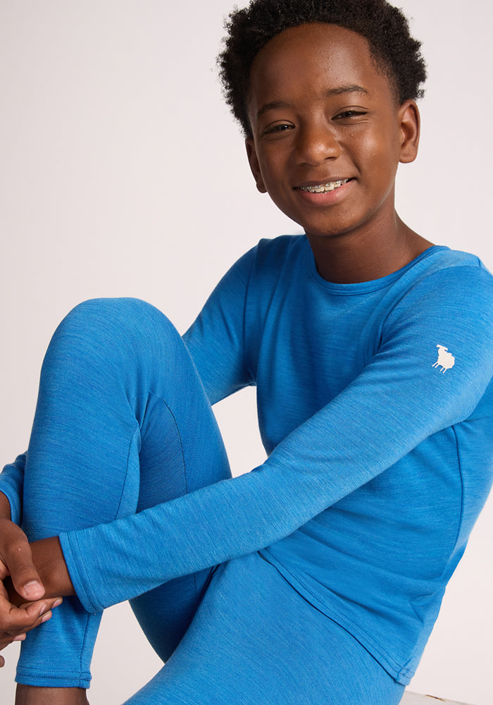 A smiling child with short curly hair wears the Woolx Kids Explorer Base Layer Top in Blue Razz, made from soft Australian Merino wool and featuring a small white sheep logo on the sleeve.