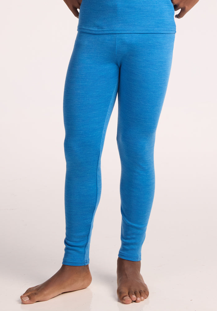 A child stands barefoot on a light surface, hands by their hips, wearing Woolx Kids Explorer Base Layer Bottoms in Blue Razz. The upper body is partially visible.