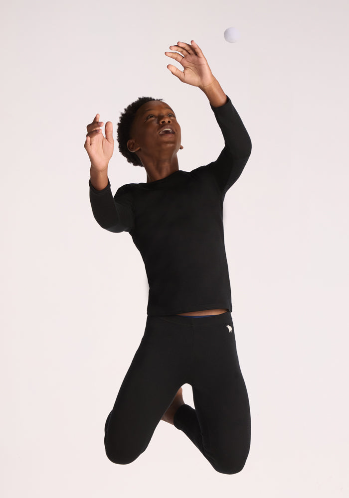A young boy in a Woolx Kids Explorer Base Layer Top - Black jumps with arms raised, reaching toward a white ball against a plain light background.