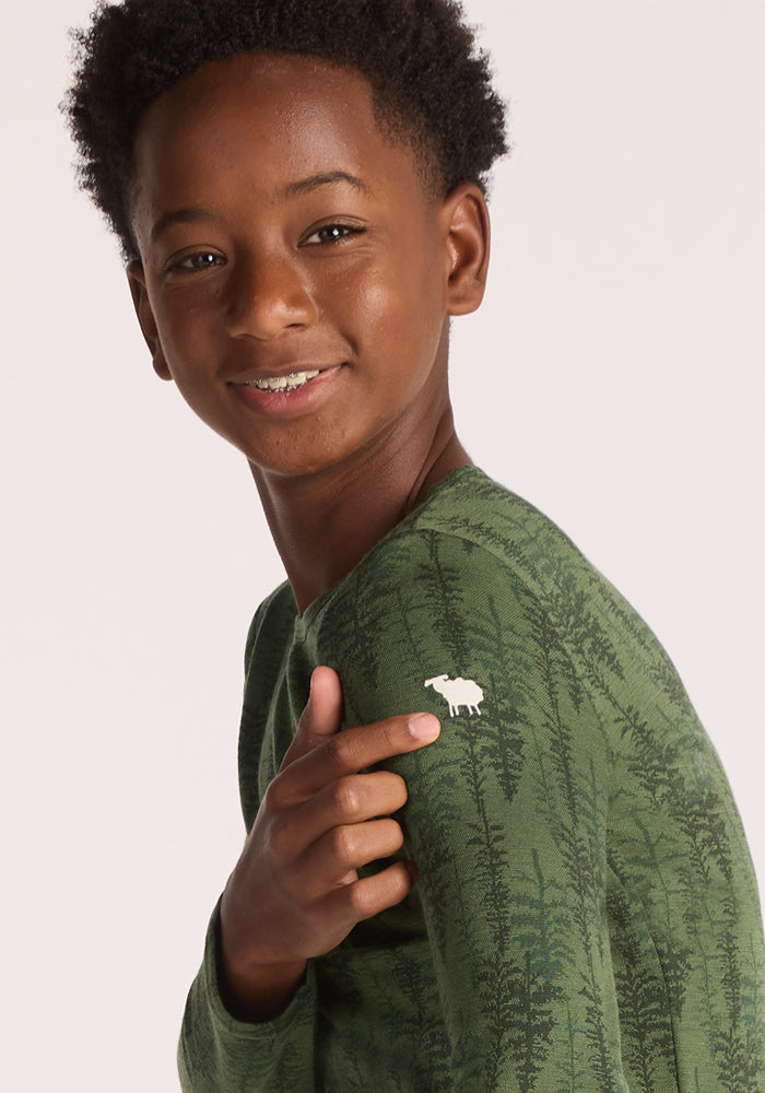 A smiling boy with short curly hair wears the Woolx Kids Explorer Base Layer Top - Forest Friends, featuring a green tree pattern and a small white moose logo on his shoulder. The background is plain and light-colored.