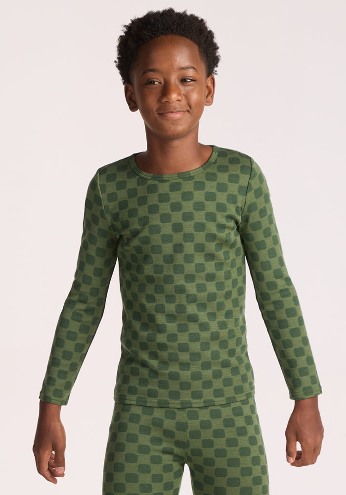 A young boy with short curly hair smiles while wearing the Woolx Kids Explorer Base Layer Top - Frog Hop, standing against a plain light background and showing off his cozy, breathable outfit.