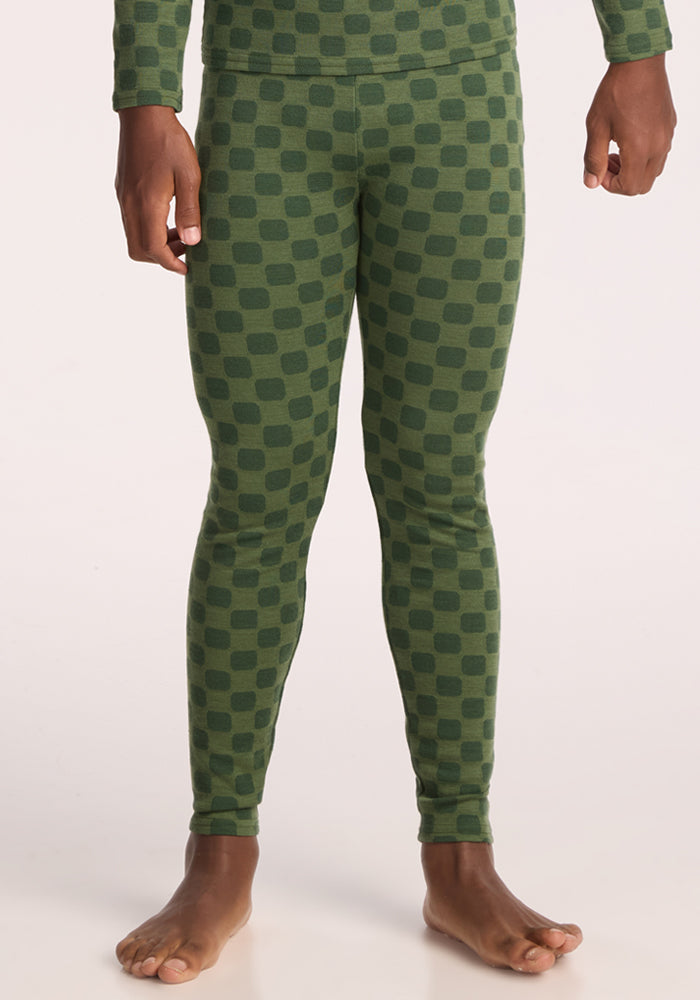 A person stands barefoot, shown from the shoulders down, wearing Woolx Kids Explorer Base Layer Bottoms - Frog Hop and a matching green checkered long-sleeve top on a light background. 