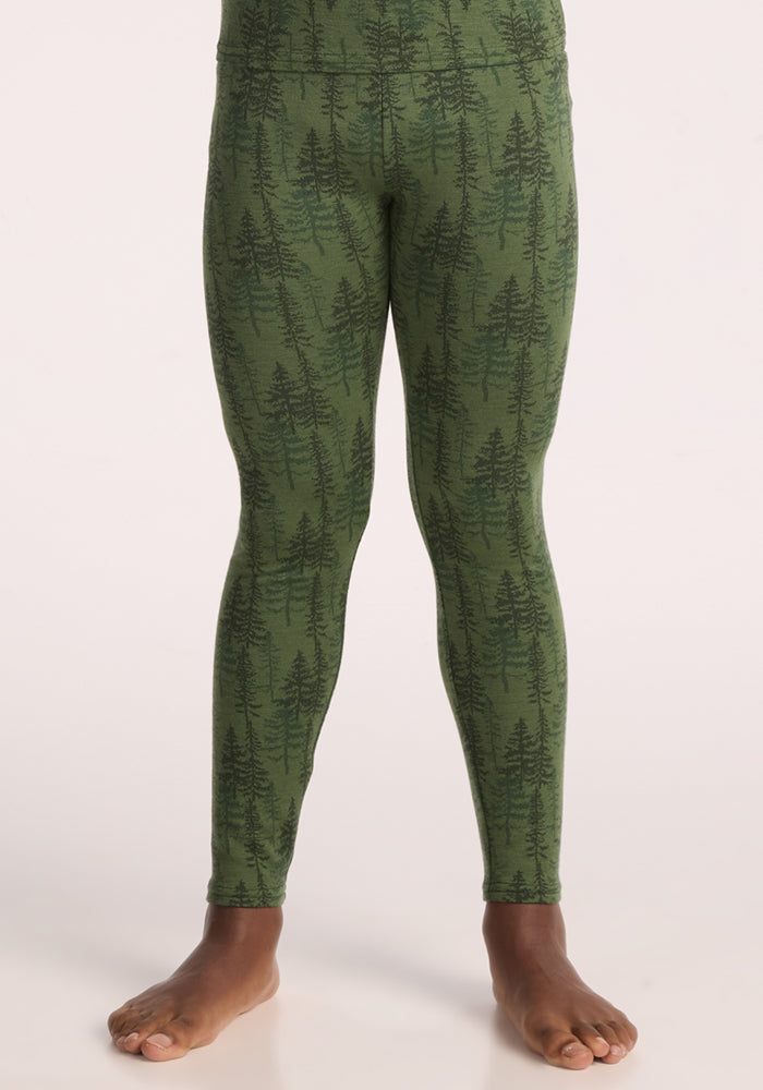 A child models Woolx Kids Explorer Base Layer Bottoms - Forest Friends, featuring a dark green tree pattern, standing barefoot against a light background. Only the lower body and legs appear to showcase these cozy merino wool leggings.