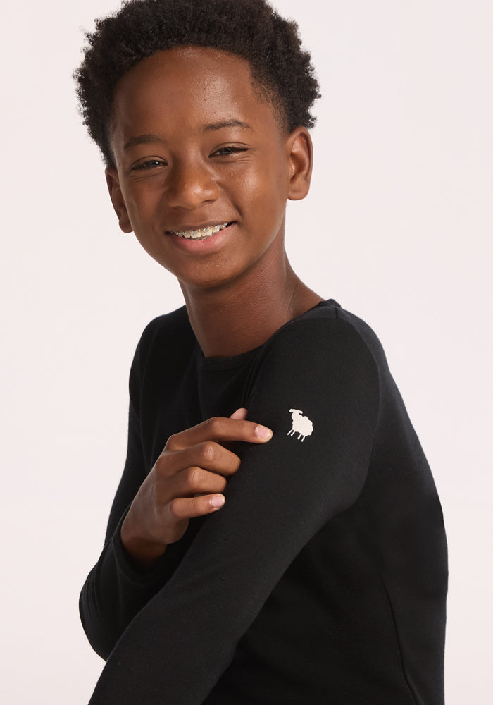 A smiling young person with short curly hair wears the Woolx Kids Explorer Base Layer Top in black, pointing to a small white sheep logo on their shoulder against a plain, light background.