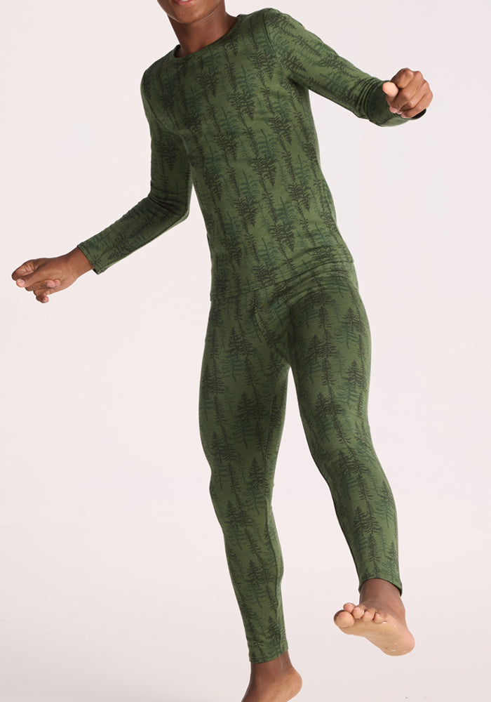 A child wears Woolx Kids Explorer Base Layer Bottoms - Forest Friends, featuring a green, full-length merino wool design with a dark tree pattern, standing barefoot against a plain light background.