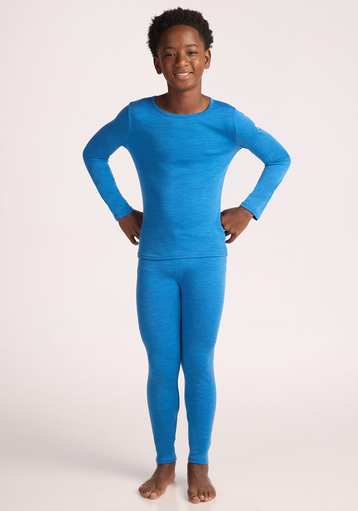 A smiling child stands barefoot with hands on hips, wearing the Woolx Kids Explorer Base Layer Top in Blue Razz, paired with matching leggings, against a plain light background.