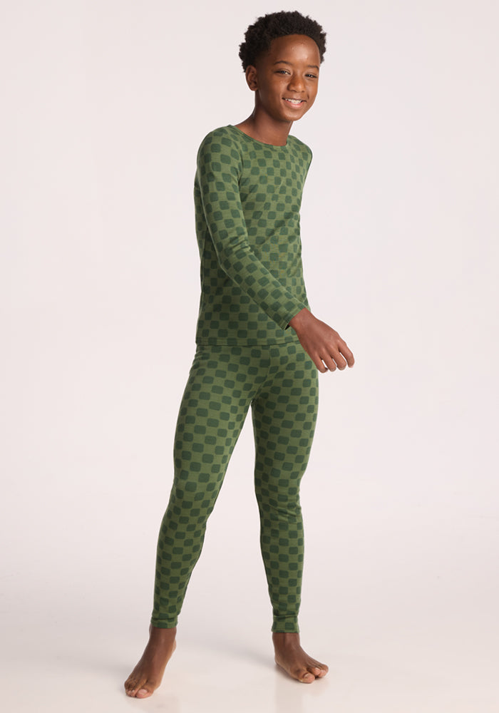 A smiling child stands barefoot on a white background, wearing Woolx Kids Explorer Base Layer Bottoms - Frog Hop, a cozy long-sleeve green pajama set with a dark green checker pattern.