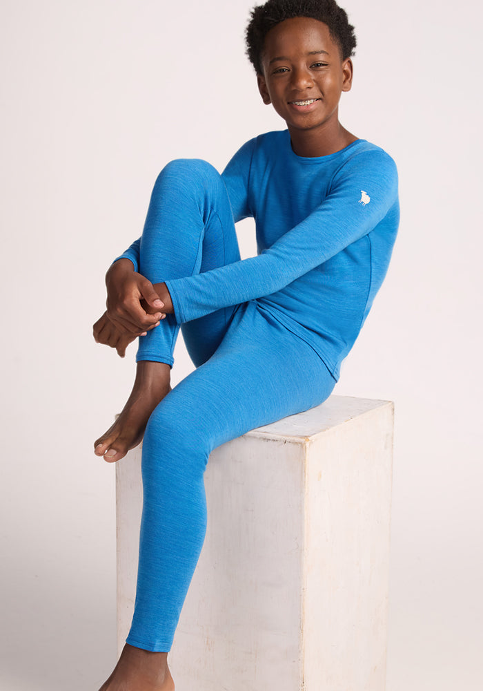 A smiling boy with short curly hair sits on a white wooden block, wearing Woolx Kids Explorer Base Layer Bottoms in Blue Razz and a matching top, one knee up and his arms resting casually on it.