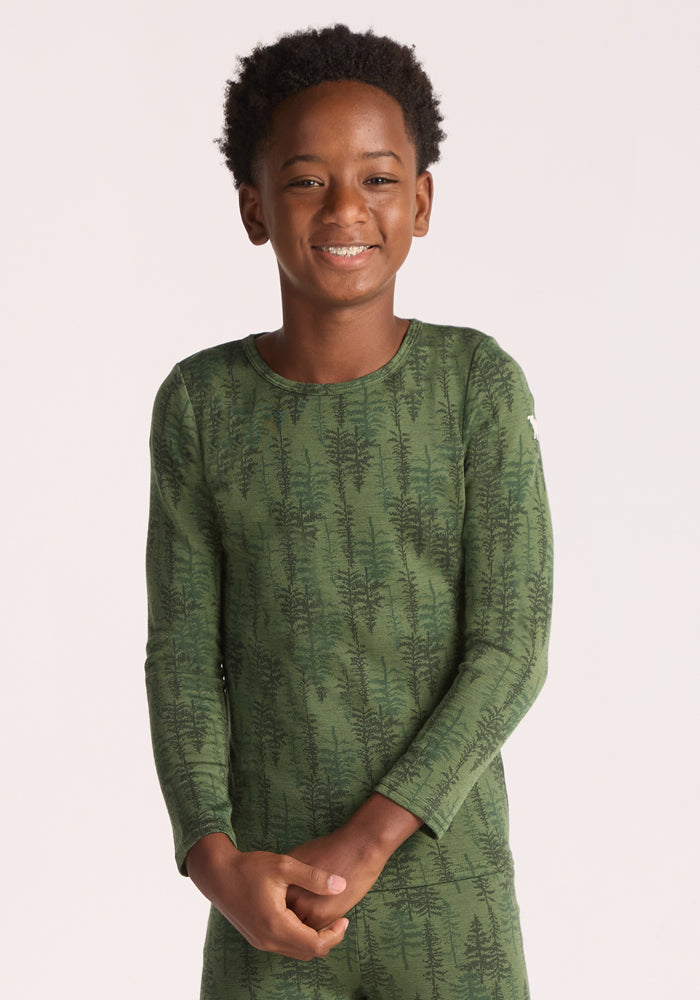 A smiling child with short curly hair wears the Woolx Kids Explorer Base Layer Top - Forest Friends, a green merino wool base layer with a subtle tree pattern, set against a plain light background. 