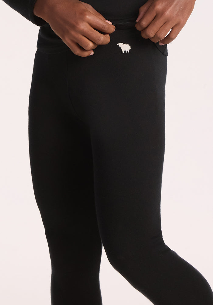 A child wearing Woolx Kids Explorer Base Layer Bottoms in black, featuring a small white sheep logo near the waistband, adjusts the waistband with both hands against a plain light-colored background.