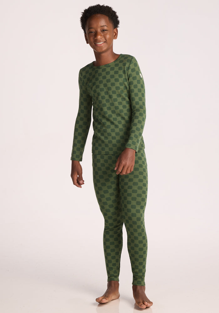 A smiling child stands barefoot on a light background, wearing the Woolx Kids Explorer Base Layer Top - Frog Hop: a breathable, soft Merino wool long-sleeve top and leggings set with a green checkerboard pattern.