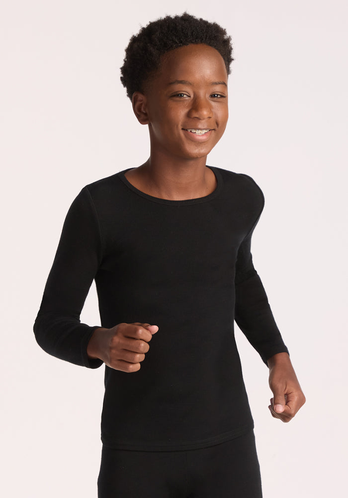 A smiling boy with short curly hair models the Woolx Kids Explorer Base Layer Top in black, made from moisture-wicking fabric. He stands against a plain white background, facing forward with arms slightly bent at his sides. 