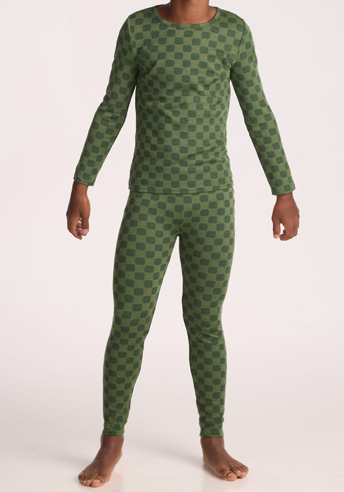 A child stands barefoot in Woolx Kids Explorer Base Layer Bottoms - Frog Hop, a merino wool set with a light and dark green checkered pattern. The matching long-sleeve top and bottoms are shown from the neck down.