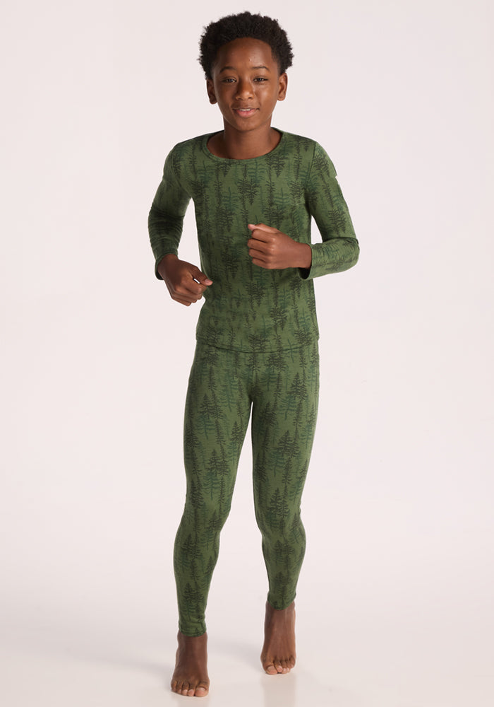 A child stands barefoot on a white background, smiling in Woolx Kids Explorer Base Layer Bottoms - Forest Friends—a cozy, stylish green merino wool baselayer featuring a dark tree pattern for warmth and comfort.