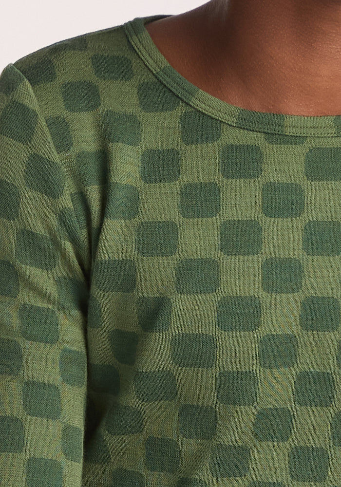 A close-up of the Woolx Kids Explorer Base Layer Top - Frog Hop in green Merino wool, featuring a geometric square pattern in dark and light green. Part of a child’s neck and shoulder shows the breathable fabric.