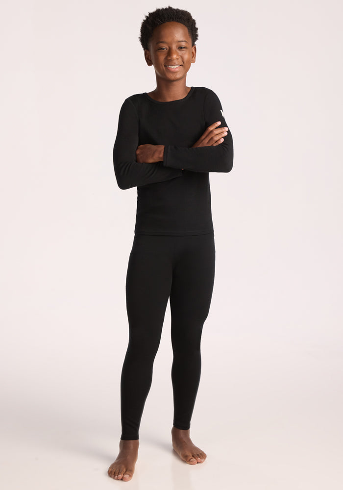 A smiling child stands barefoot with arms crossed, wearing Woolx Kids Explorer Base Layer Bottoms in black and a matching merino wool top, set against a plain light background.
