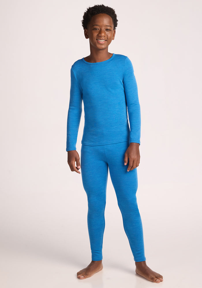 A smiling child stands barefoot on a neutral background, wearing Woolx Kids Explorer Base Layer Bottoms in Blue Razz—a cozy, long-sleeve toddler thermal ideal for cool days.