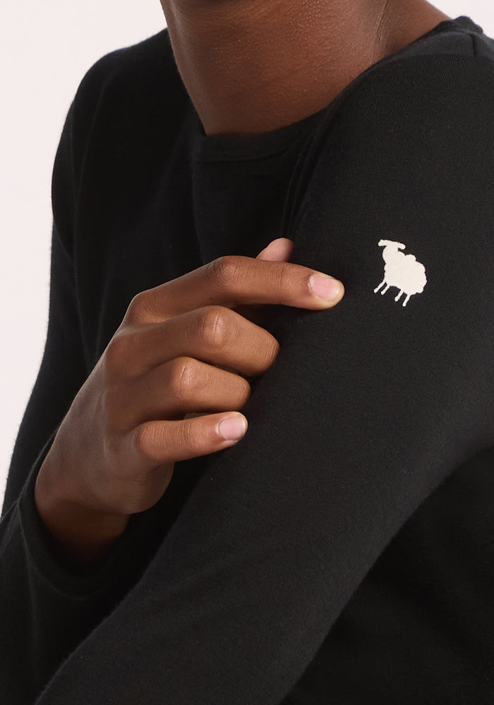 A child wearing the Woolx Kids Explorer Base Layer Top in black points to a small white sheep graphic on the upper arm. The background is plain and light-colored.