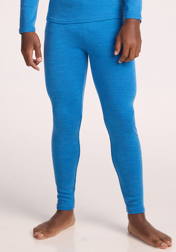 A person stands barefoot in Woolx Kids Explorer Base Layer Bottoms - Blue Razz and a matching blue long-sleeve top, shown from shoulders to feet against a plain light background.