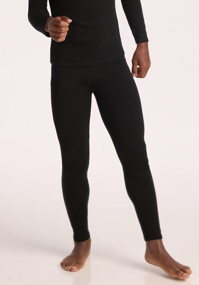 A person stands barefoot, wearing fitted black long-sleeve top and Woolx Kids Explorer Base Layer Bottoms in black, with one arm bent and the other hanging naturally. The photo is cropped above the shoulders.