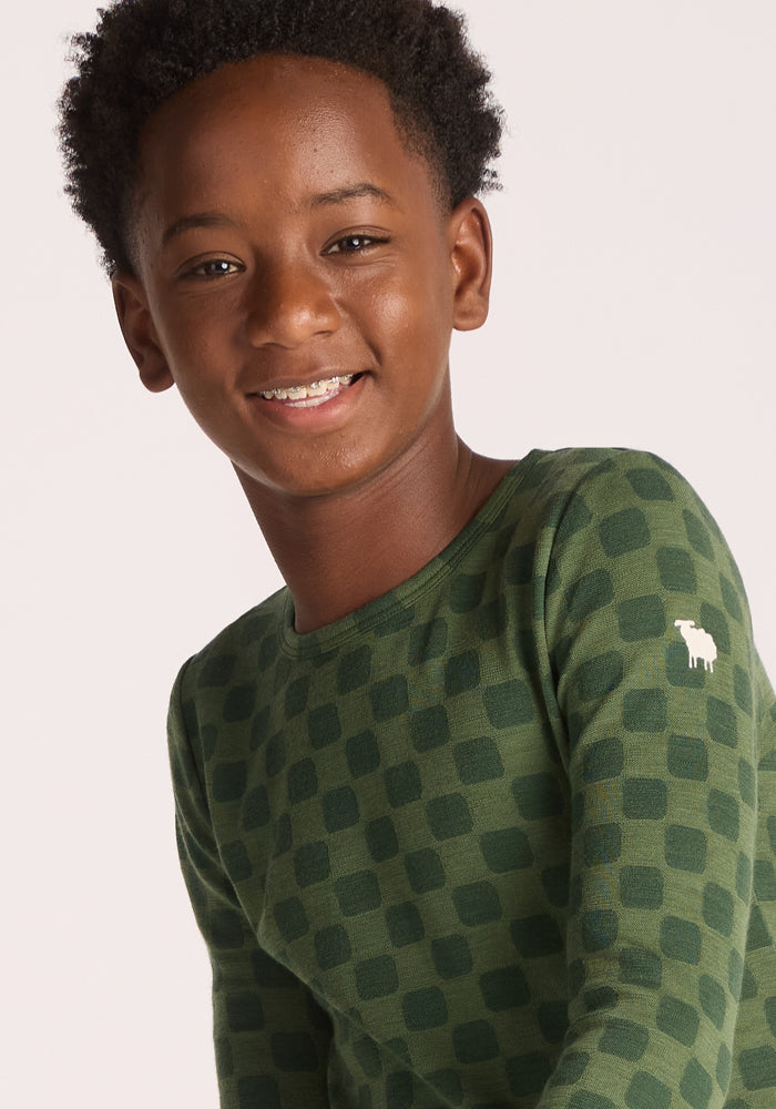 A smiling boy with short curly hair wears the Woolx Kids Explorer Base Layer Top - Frog Hop, a green checkered kids' wool thermal featuring a small white animal logo on the shoulder. He faces slightly to the side against a plain light background.