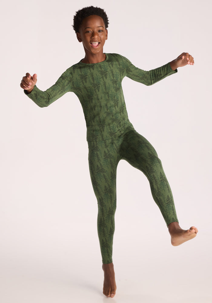 A smiling child in the Woolx Kids Explorer Base Layer Top – Forest Friends, a soft Australian Merino wool long-sleeve with a tree print, poses joyfully on one foot against a plain light background.