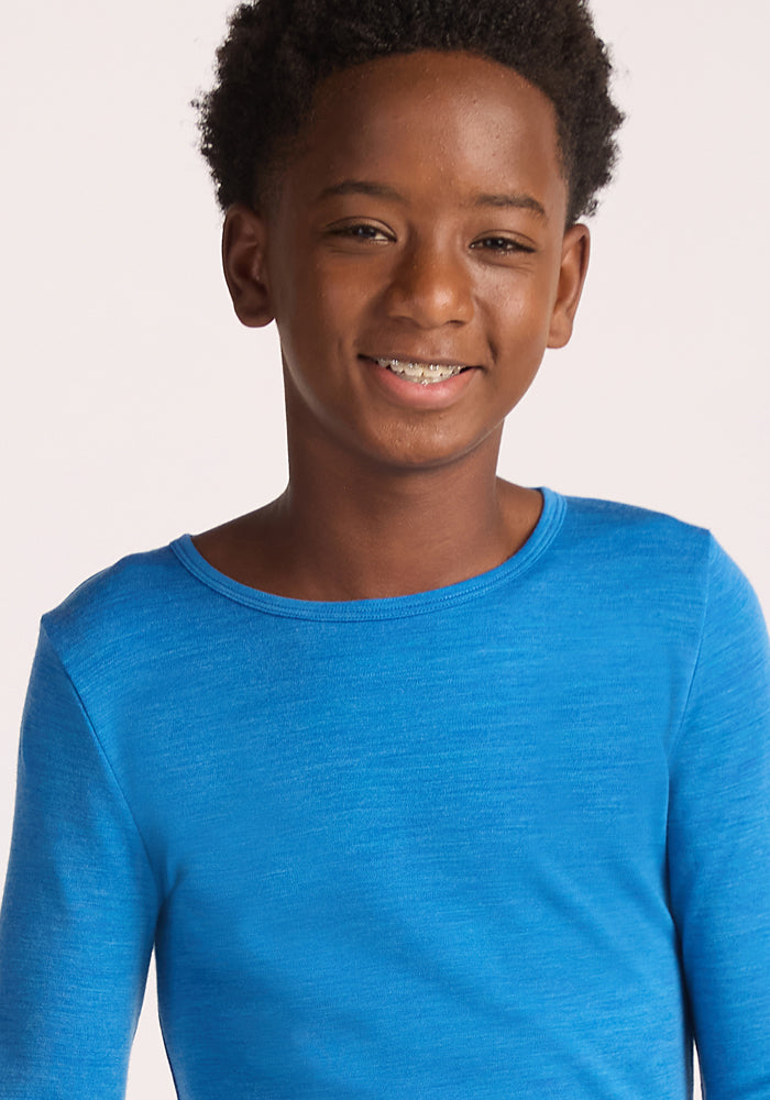A smiling boy with short curly hair wears the Woolx Kids Explorer Base Layer Top in Blue Razz, standing against a plain light background.