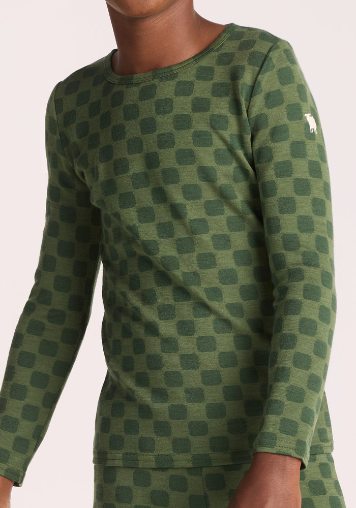 A child wears the Woolx Kids Explorer Base Layer Top - Frog Hop, a breathable, long-sleeve green Merino wool shirt featuring a checkerboard pattern and a small white logo on the upper arm.