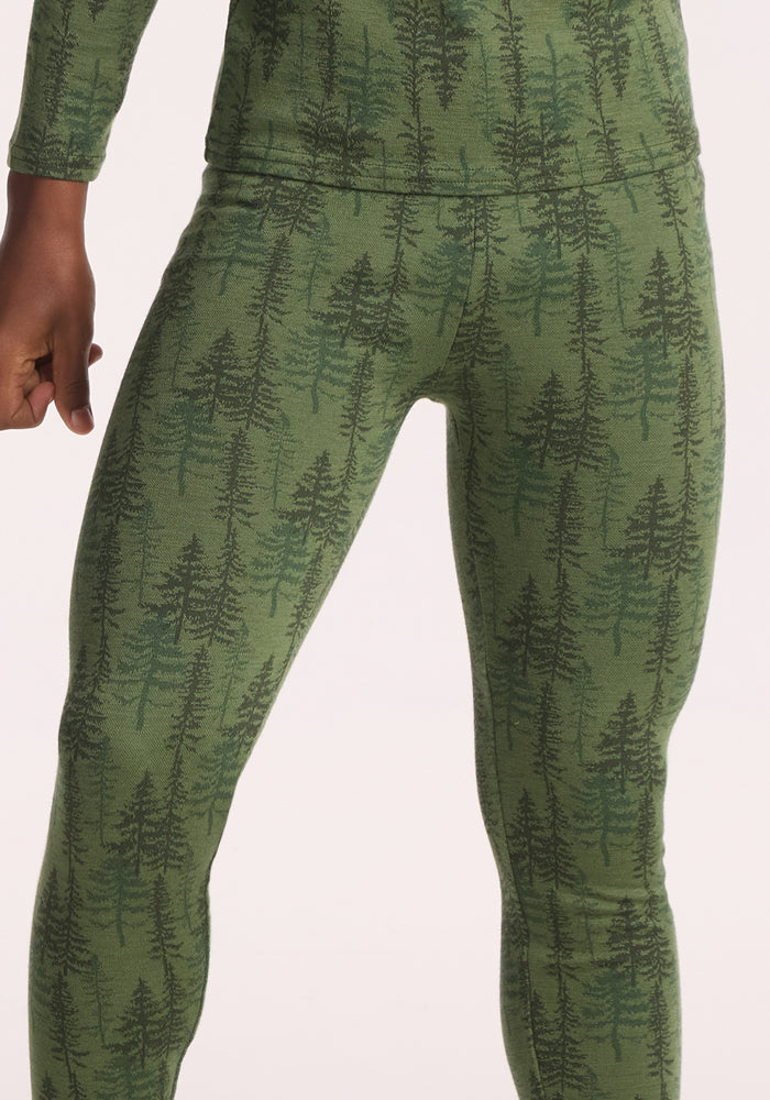 A child models Woolx Kids Explorer Base Layer Bottoms - Forest Friends, in green with a dark tree pattern, shown waist to mid-thigh with one arm bent and fist visible, against a white background.