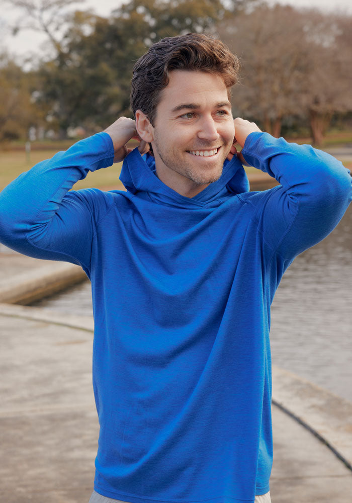 A man smiles as he pulls up the hood of his Woolx Base Camp Hoodie in Glacial Blue while standing outdoors near a curved walkway and water, with trees and grass in the background.