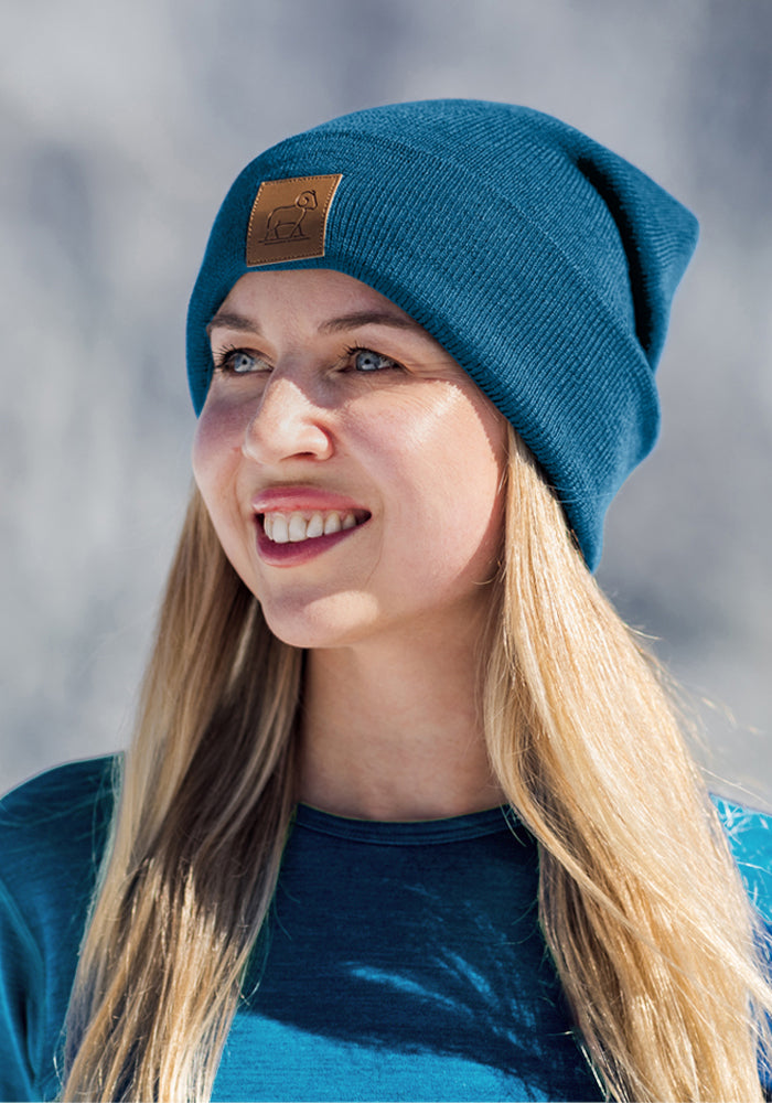 Smiling woman with long blonde hair in a Woolx Baylor Beanie - Blue Pine and matching shirt, outside in winter.