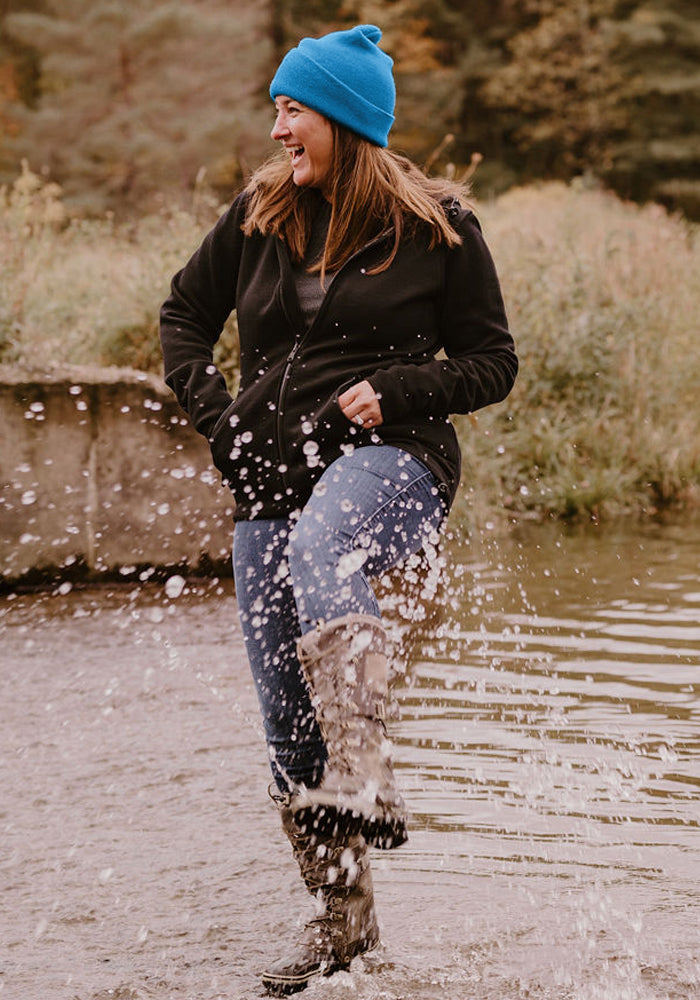 Woman in a Woolx Baylor Beanie - Blue Razz smiles, splashing water in her black jacket and boots by the stream.