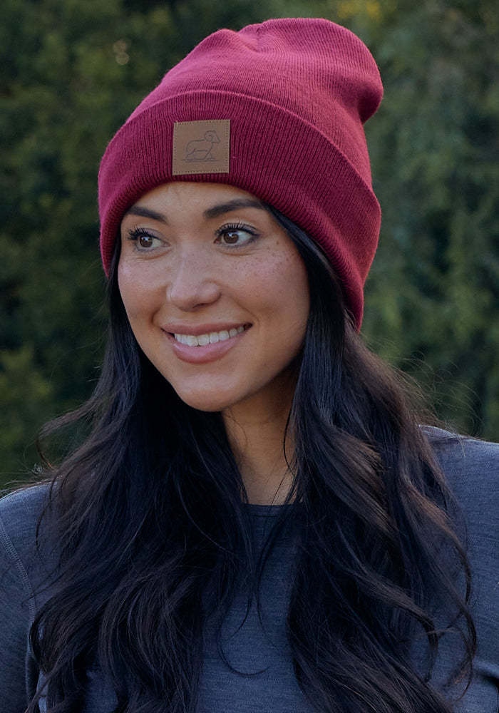A woman with long dark hair smiles while wearing the Woolx Baylor Beanie in Sweet Cream and a dark gray top, standing outdoors with a blurred green background.