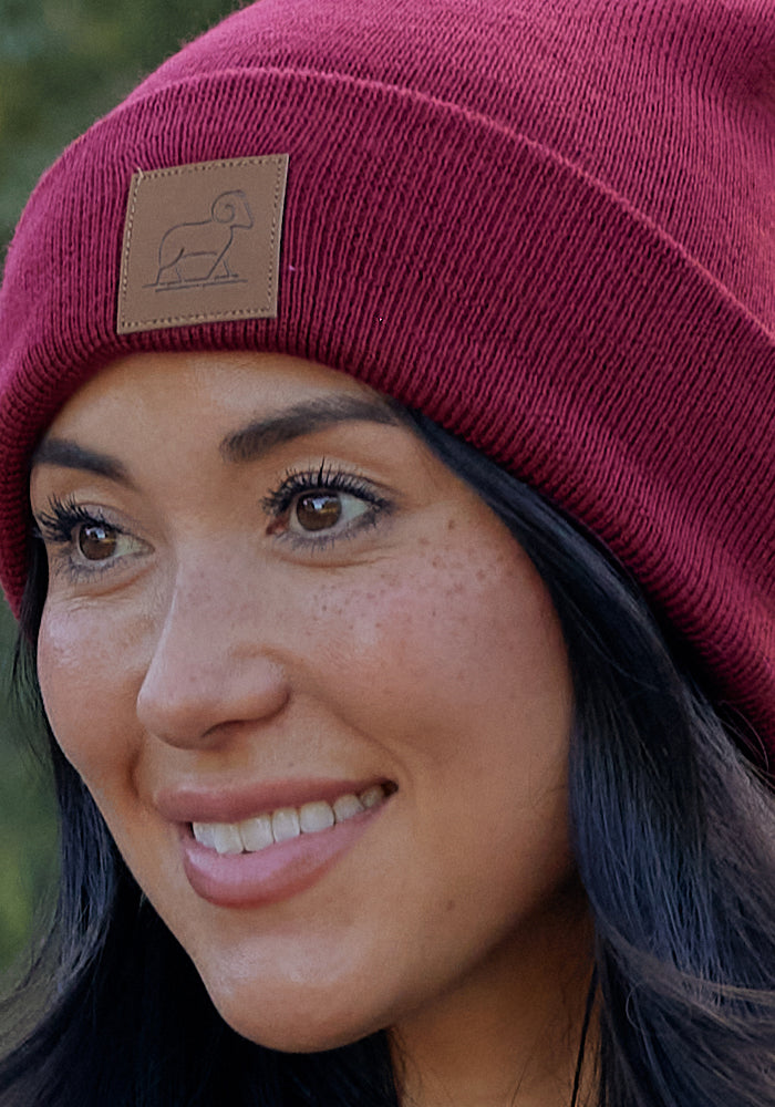 A woman with long dark hair smiles while wearing the Woolx Baylor Beanie in Toasted Garnet, featuring a brown patch with a simple ram logo—making it the perfect warm winter hat.