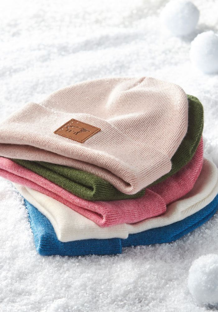 A stack of pastel knit beanies, including the Woolx Kids Baylor Beanie in Pinky Promise, rests on snowy ground with snowballs in the background. The top beanie has a brown leather patch for a cozy winter look.