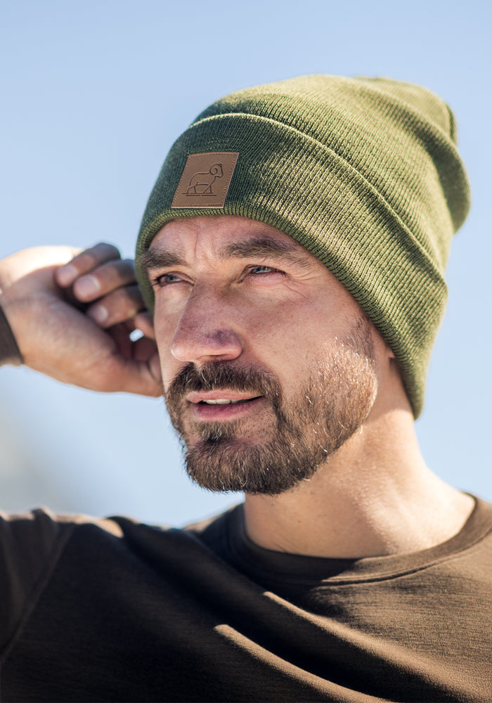 A man with a short beard, wearing the Woolx Baylor Beanie in Winter Moss and a brown shirt, gazes into the distance beneath a clear blue sky.