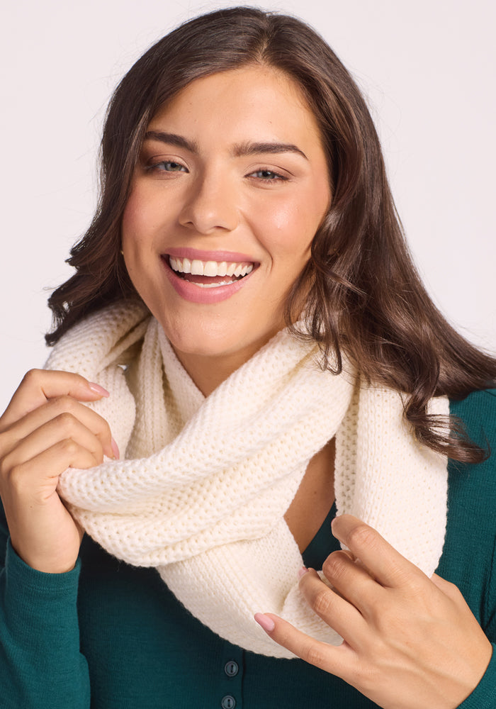 A smiling woman with long brown hair wears the Woolx Noelle Infinity Scarf in Buttercream as a cozy winter accessory over a green top, holding the soft merino wool scarf with both hands against a plain light background.