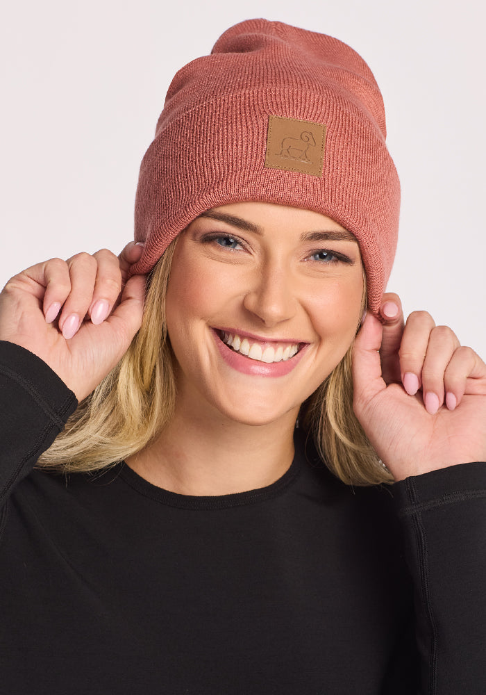 A smiling woman with long blonde hair and natural makeup poses in front of a white background wearing the Woolx Baylor Beanie in Brick Dust and a black long-sleeve shirt, holding the sides of her hat.