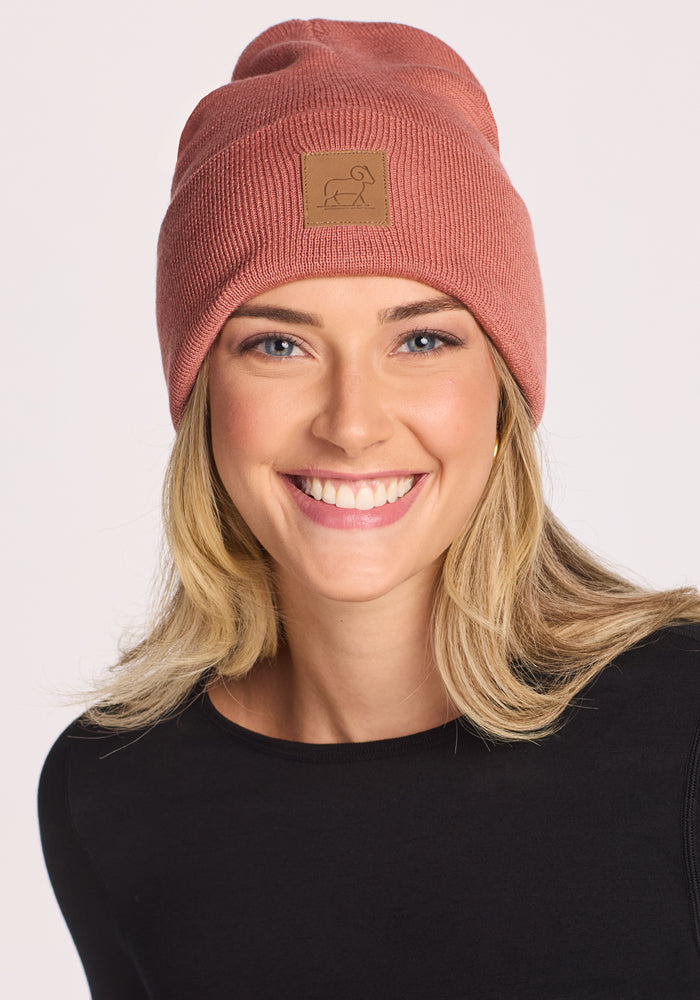 A smiling woman with blonde hair wears the Woolx Baylor Beanie in Brick Dust and a black top, posing against a plain light background.