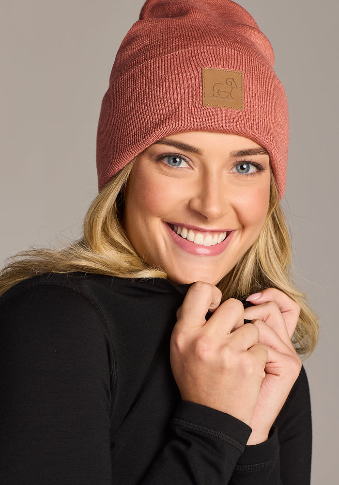 A smiling woman wearing the Woolx Baylor Beanie in Brick Dust and a black long-sleeve top, holding the collar close to her face against a neutral background.
