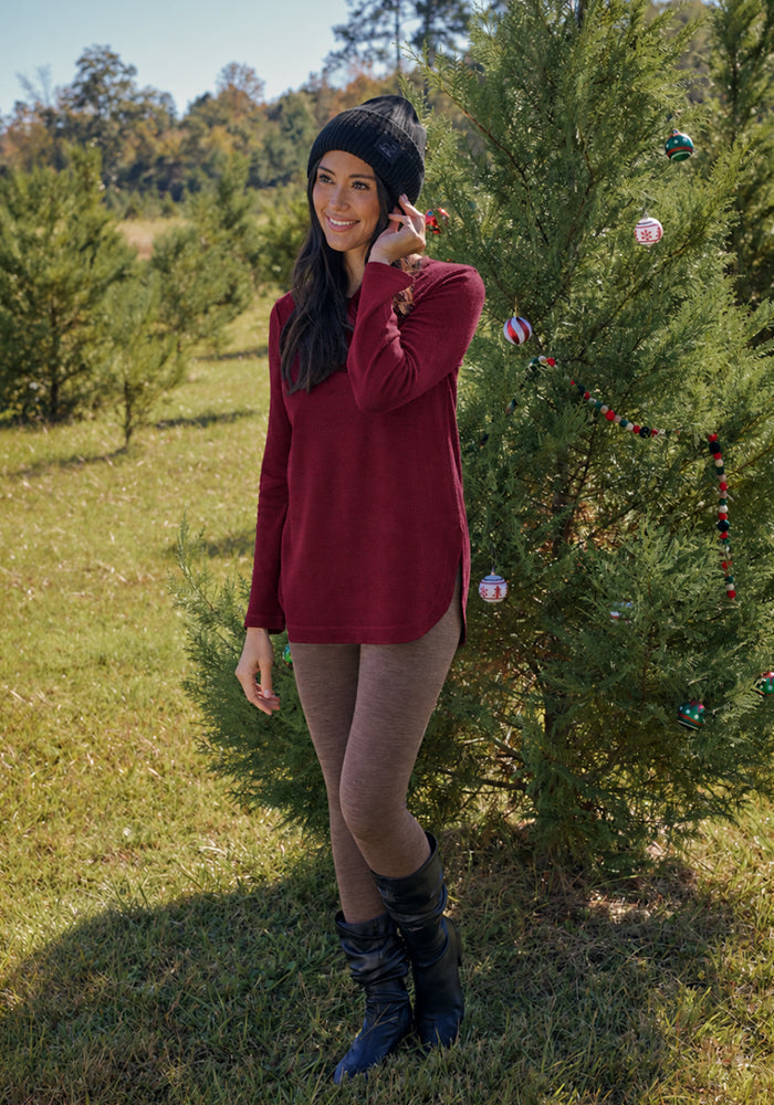 Woman in Woolx Blair Tunic - Beet Red and beanie smiles by a festive tree—ideal for chic, lightweight travel.