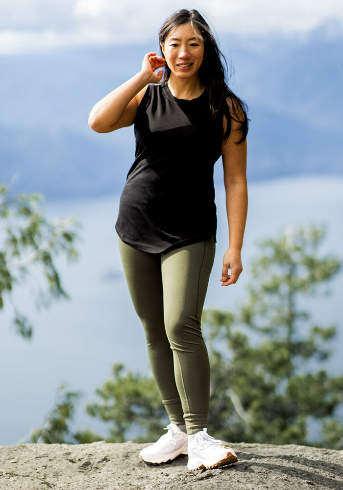 A woman wearing the Woolx Bella Tunic Top in black, paired with olive leggings and white sneakers, stands on a rock outdoors by blurred trees and water, smiling as she touches her hair.