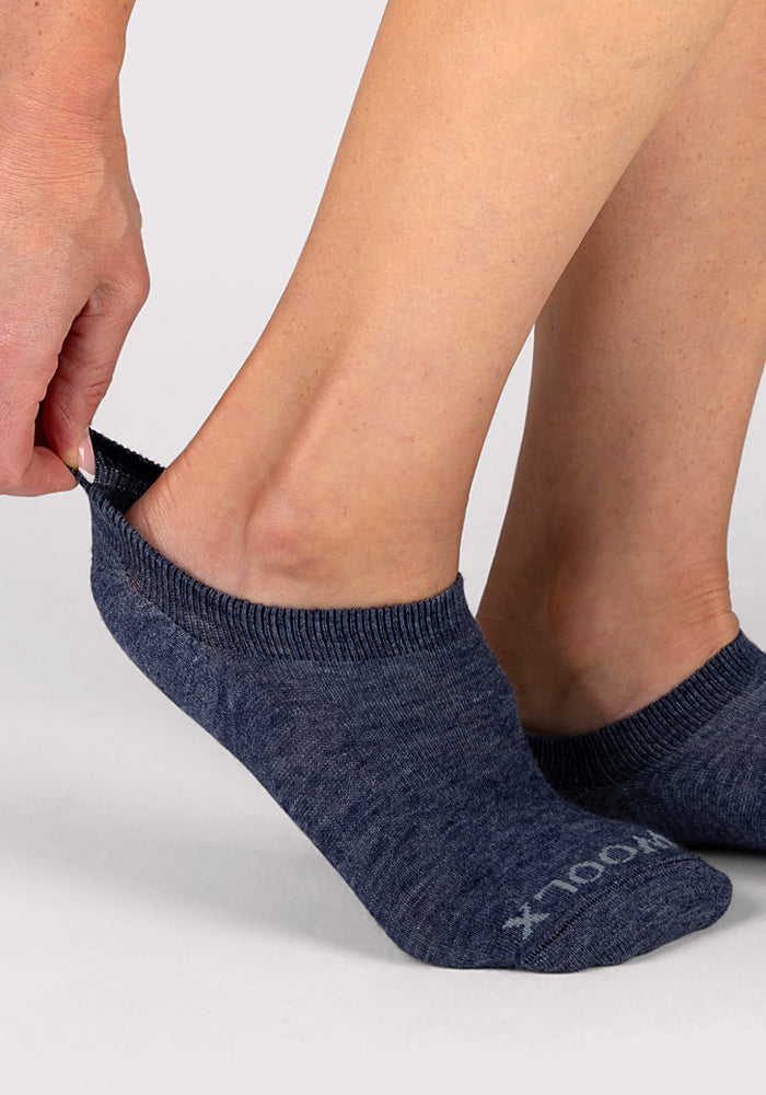 A person puts on a low-cut Woolx Womens Daisy Ped Sock in Blue Jean Dream, featuring “WOOL” on the toe. The no-show sock covers the foot and ankle against a light gray background.